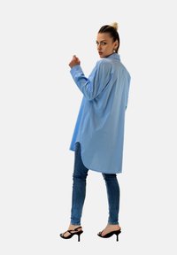 Camicia a maniche lunghe azzurro chiaro con vestibilità rilassata, orlo curvo, indossata su jeans di denim scuri e tacchi neri a strappo. Tessuto dalla trama liscia.