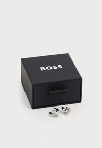 Pendientes en tono plateado exhibidos en una caja negra de BOSS. Los pendientes presentan un acabado brillante y un diseño adornado. La caja tiene un cierre de lengüeta.