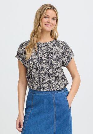 Jeune femme aux longs cheveux blonds portant un haut à fleurs noir et blanc et une jupe en jean bleue, souriante avec une main dans la poche.