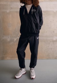 Svart vindjacka med vit logotyp, i kombination med matchande byxor. Skorna är grå och vita med röda accenter. Slät textur, modern design.
