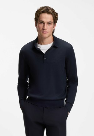 Ung mand iført en marineblå langærmet poloshirt over en hvid t-shirt, stående med hænderne i lommerne mod en ensfarvet hvid baggrund.