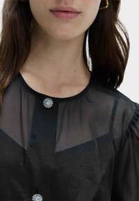 Zwarte doorschijnende blouse met een ronde halslijn, voorzien van een delicate zilveren bloempjesknop op de kraag en subtiele pofmouwen.