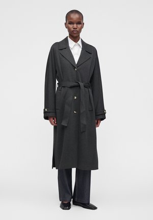 Personne portant un long manteau gris foncé ceinturé sur une chemise blanche à col et un pantalon large foncé, debout devant un fond clair uni.