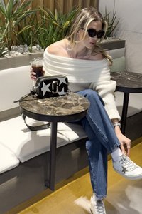 Off-white, ribbed off-shoulder neule; farkut; valkoiset lenkkikengät mustalla tähdellä; musta käsilaukku valkoisilla tähdillä; pyöreä marmoripöytä.