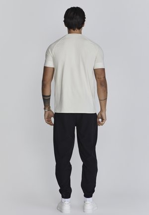 Světlé béžové tričko s krátkým rukávem, běžného střihu, v kombinaci s elastickými černými tepláky a bílými teniskami, pohled zepředu.