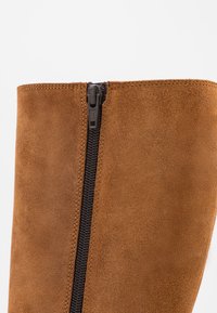 Brauner Wildlederstiefel mit seitlichem Reißverschluss, glatter Textur und anliegendem Schaft. Der Reißverschluss ist schwarz mit sichtbaren Zähnen und einem Ziehschieber.