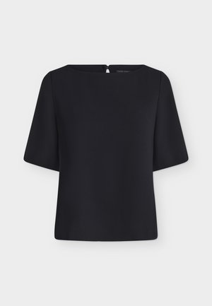 Donna con top nero a maniche corte, pantaloni marroni a gamba larga, sandali neri con tacco, che tiene una borsa nera con frange su uno sfondo semplice.