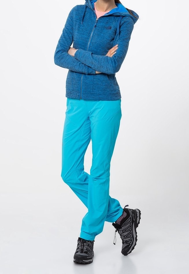 Personne portant un sweat à capuche bleu zippé, un pantalon turquoise vif et des chaussures de randonnée noires, debout les bras croisés et une jambe légèrement pliée.