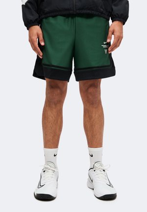 Personne portant un short de basketball Milwaukee Bucks vert foncé, des chaussettes blanches Nike et des chaussures de sport blanches avec des touches noires.