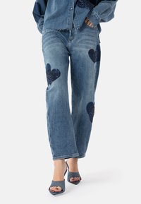 Jeans a gamba larga di un blu chiaro con toppe a forma di cuore glitterate di un blu scuro su entrambe le gambe; abbinati a sandali con tacco aperti di un blu scuro.