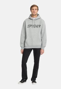 Felpa grigia con il logo "SPIDER" in nero sul petto, abbinata a pantaloni neri e sneakers nere. Il tessuto appare morbido, vestibilità classica.