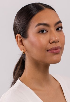 Goldene Creolen mit einem gedrehten Design. Das Modell hat dunkles Haar, das zu einem niedrigen Pferdeschwanz frisiert ist und trägt ein weißes Oberteil mit V-Ausschnitt.