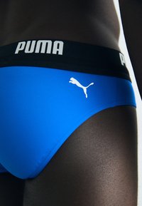 Kék úszónadrág fekete derékrésszel, melyen fehér "PUMA" logó és egy kis fehér ugró puma embléma található az oldalon.