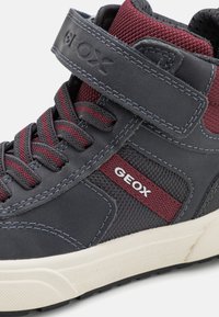 Korkeat tennarit harmaasta synteettisestä materiaalista, jossa on viininpunaisia korostuksia, teksturoitu verkko, nauhakiinnitys ja tarranauha. GEox-logo näkyvissä sivulla.