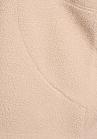 Beige fleece stof met een zachte, gestructureerde oppervlakte, voorzien van een zijzak en gladde naden. Het materiaal lijkt warm en fluffig.