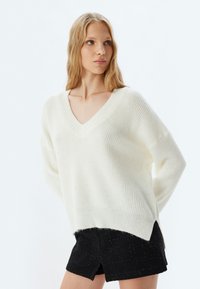 Cremefarbener Pullover mit V-Ausschnitt, geripptem Material, überschnittenen Schultern und einem leicht verlängerten Rückensaum, kombiniert mit strukturierten schwarzen Shorts.