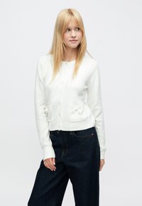 Fehér kötött kardigán kerek nyakkivágással, gombos záródással és két elöl található, masnival díszített zsebbel. Sötétkék farmer széles szárú nadrággal párosítva.