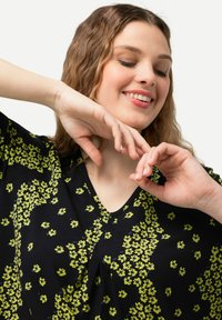 Černý top s výstřihem do V, zdobený živou žlutou květinovou vzorovanou. Měkká tkanina s uvolněným střihem a plynulými rukávy.