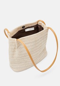 Woven straw tote bag w zewnątrz w jasnym beżu z brązowymi skórzanymi uchwytami. Wnętrze w kolorze brązowym z metką logo. Otwarte z góry z zapięciem na magnes.