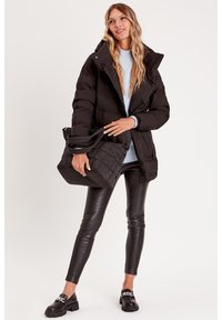 Schwarze Daunenjacke mit Kapuze, kombiniert mit einem hellblauen Pullover, schwarzen Lederhosen und einer gesteppten schwarzen Tasche. Klobige schwarze Schuhe.