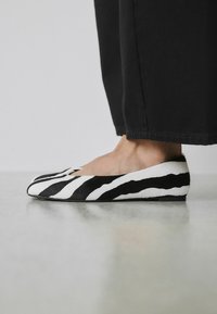 Chaussures plates à rayures noires et blanches en matériau texturé, avec un bout arrondi et un profil bas. Fond neutre.