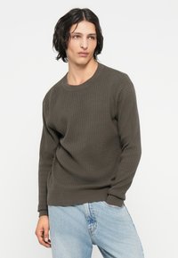 Ciemnooliwkowy, prążkowany sweter z okrągłym dekoltem i długim rękawem, w zestawie z jasnoniebieskimi dżinsami.