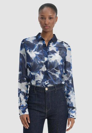 Donna che indossa una camicia aderente a maniche lunghe con bottoni, con motivi astratti blu e bianchi, e jeans in denim scuro a vita alta.