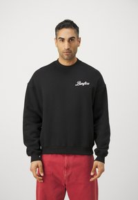 Sudadera negra con cuello redondo y mangas largas, que presenta el texto "Benfica" en blanco en el pecho, combinada con pantalones rojos.
