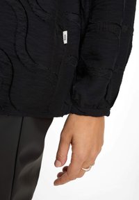 Black textured long-sleeve top with a wave pattern, featuring a label on the side. Hand visible, wearing a silver ring.