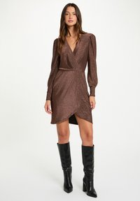 Abito avvolgente metallizzato marrone con maniche lunghe a sbuffo, lunghezza a metà coscia, caratterizzato da una scollatura a V e un dettaglio arricciato sul lato. Abbinato a stivali neri al ginocchio.