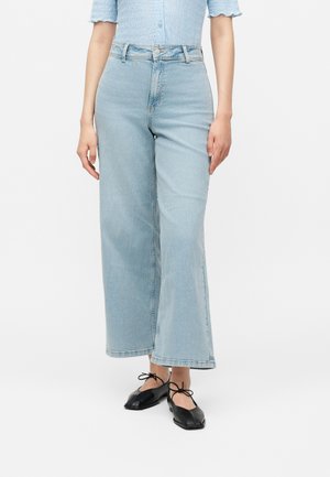 Persona con vaqueros de tiro alto y pierna ancha en azul claro, camiseta texturizada azul claro y bailarinas negras con lazadas en el tobillo, de pie sobre fondo blanco.