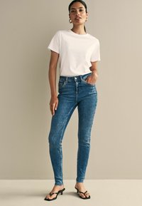 Plave skinny traperice s visokim strukom, u kombinaciji s jednostavnom bijelom majicom. Glatka tekstura i minimalni detalji. Crne sandale na visoku petu.