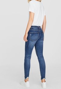 Smala jeans i mörkblå denim med fransade fållar, hög midja och dekorativ sömnad på bakfickorna. Matchas med en vit t-shirt.