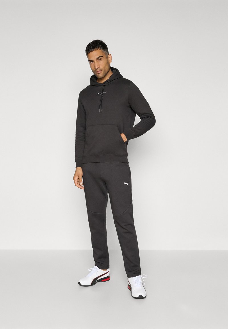 Sudadera negra con cordones y bolsillo frontal; pantalones de chándal a juego con paneles texturizados; modelo que lleva zapatillas blancas con acentos rojos y negros.