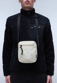 Beige crossbody bag with a zippered closure, featuring a circular logo and graphics. Worn over a black fleece jacket with a zippered pocket.