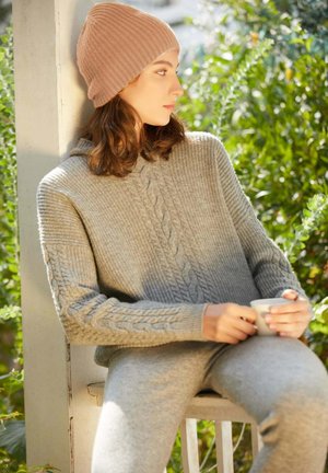 Grauer Kabelstrickpullover mit Kapuze, gerippten Ärmeln und passenden grauen Hosen. Das Model hält einen weißen Becher und trägt eine hellbeige Mütze.