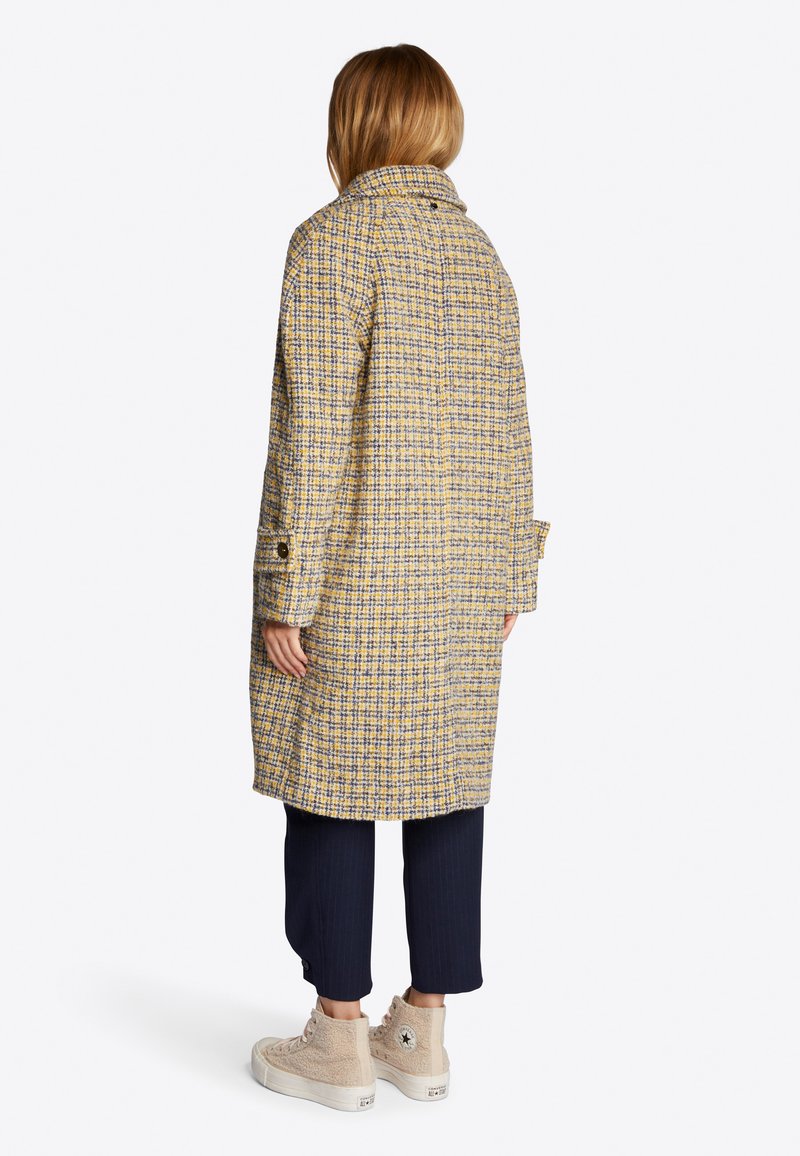 Persona vistiendo un abrigo largo a cuadros amarillo y gris, pantalones cortos navy y zapatillas altas beige, de pie de espaldas sobre un fondo blanco.