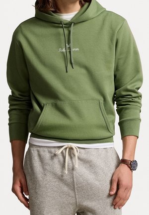 Personne portant un sweat à capuche vert avec poche avant, un sous-vêtement blanc, un pantalon de survêtement gris avec cordon de serrage et une montre-bracelet au bras gauche.