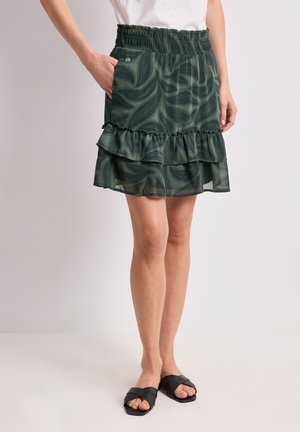 Jupe à volants à motif vert avec taille élastique et poche latérale portée avec un haut blanc et des sandales noires à enfiler sur une personne debout.