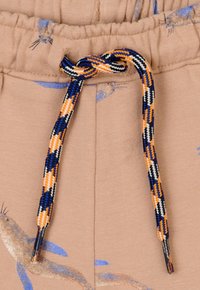 Beige sweatpants hebben een trekkoord met blauwe, oranje, zwarte en witte patronen, en kleine dierenafbeeldingen gedrukt op de stof.
