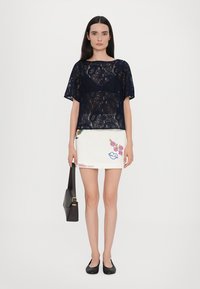 Navy lace short-sleeve top with a fitted white skirt featuring colorful floral embroidery and designs, completed with black flat shoes and a bag.