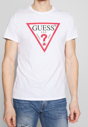 Biały bawełniany T-shirt z czerwonym trójkątnym logo z napisem "GUESS" oraz dodatkowym tekstem. Krótkie rękawy i okrągły dekolt. Noszony z niebieskimi jeansami.