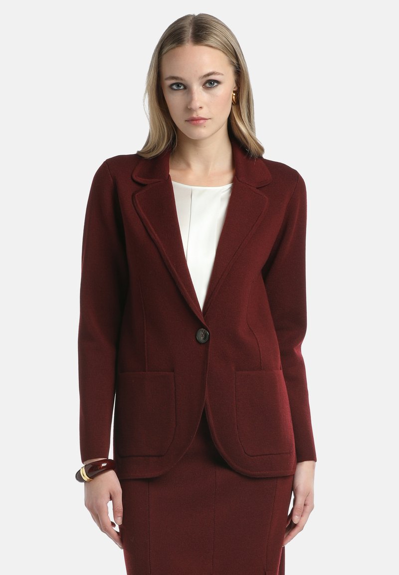Woman wearing a tailored burgundy blazer with front pockets and a button, paired with a matching skirt and white inner blouse.