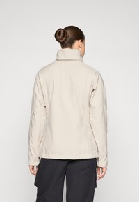 Veste en softshell beige avec un col haut, des coutures visibles et une coupe ajustée, portée sur un pantalon cargo sombre, vue de dos.