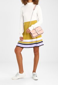 Maglietta bianca a maniche lunghe abbinata a una gonna gialla a pieghe con strisce viola e nere. Accentuata da una borsa rosa e sneaker bianche.