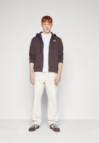 Sudadera con cremallera marrón y bolsillos frontales, llevada sobre una camiseta blanca. Pantalones color crema y zapatillas marrones con cordones blancos.