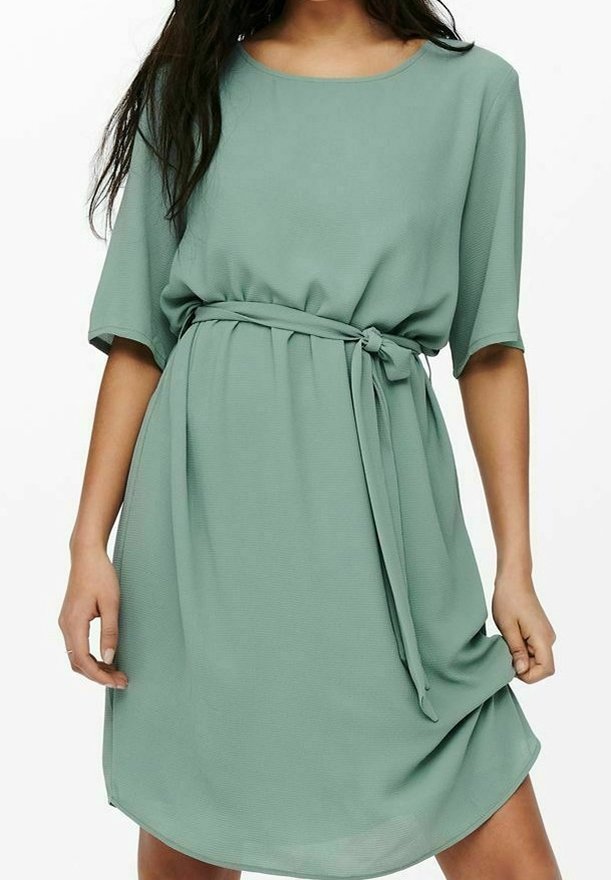 Femme portant une robe ample jusqu'aux genoux, vert sauge, avec des manches courtes et une ceinture nouée à la taille, tenant l'ourlet de la robe.