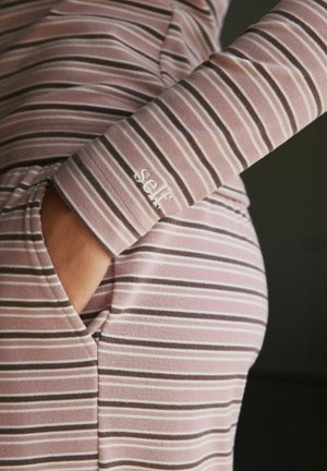 Person trägt ein pink-braun gestreiftes Outfit, eine Hand in der Tasche, Manschette mit dem aufgestickten Wort „self“.