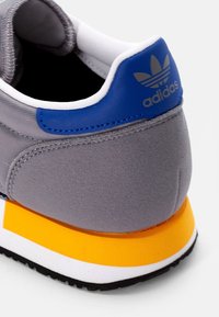 Zadní pohled na šedou sneaker od Adidas s modrým patním štítkem, žlutou mezipodešví, bílým lemováním a černou podešv�í na bílém pozadí.