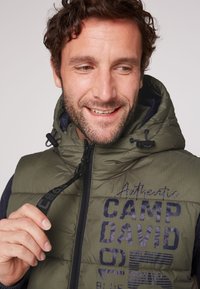 Gilet imbottito verde con cappuccio, dotato di chiusura con zip e dettagli in navy. "CAMP DAVID Autentico" stampato in blu scuro sul davanti.